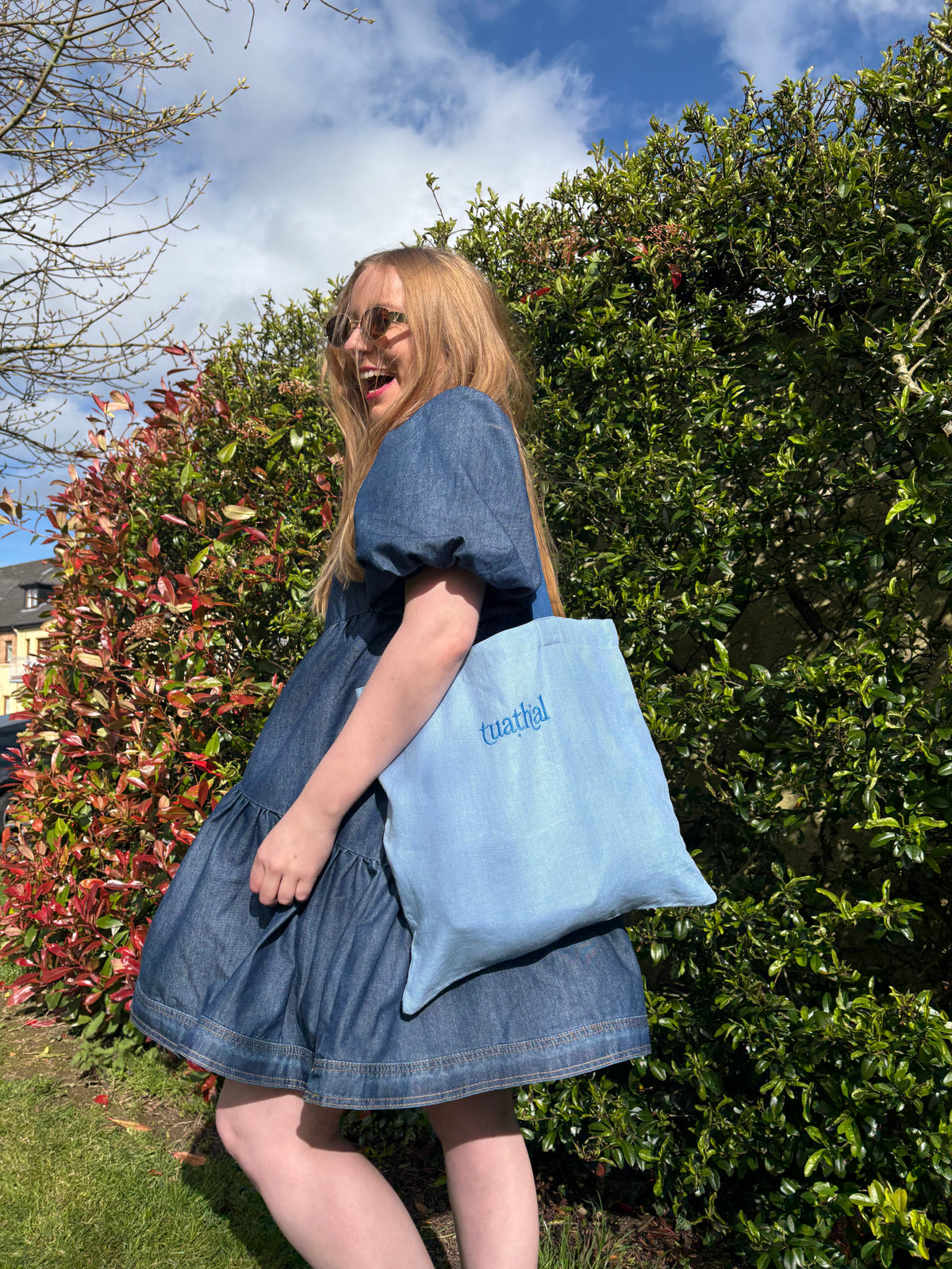 Light blue linen tote bag with Tuathal embroidered in blue, carried on the shoulder by a woman in a denim dress